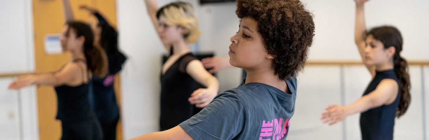 A dancer is wearing a grey t-shirt with The Place logo on the back. They are in a traditional ballet position with one arm reaching to the ceiling and the other outstretched to the side. There head is pointing diagonally as they look towards the arm to the side.