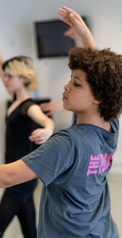 A dancer is wearing a grey t-shirt with The Place logo on the back. They are in a traditional ballet position with one arm reaching to the ceiling and the other outstretched to the side. There head is pointing diagonally as they look towards the arm to the side.