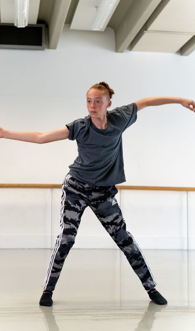 a dancer wearing a grey t-shirt and patterned grey and black trousers has one leg bent slightly and the other is stretched to the side. Their arms are curved in a wave like position out to the side and they are looking to the corner of the dance studio.