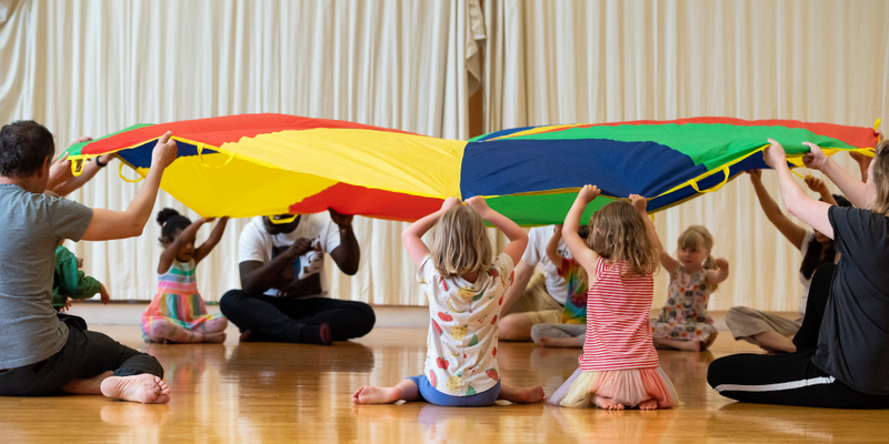 A group of children sitting in a circle on the floor holding up a parachute.