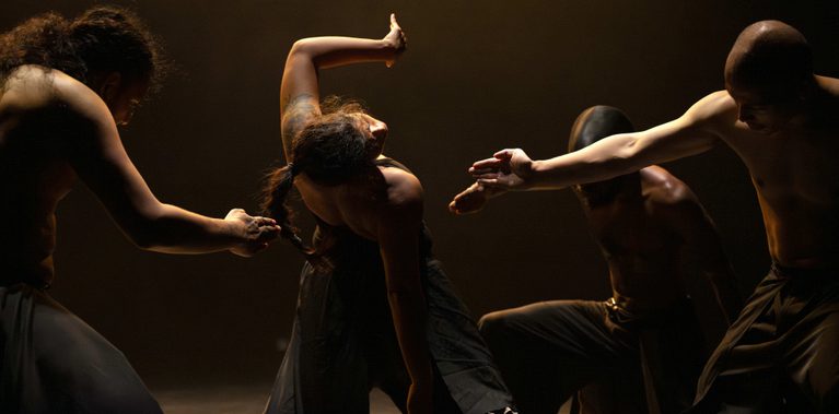 A group of people, lit by subtle light, wearing black trousers, some some shirtless, others in black tops. One person in the centre, seen from behind, raises one arm and leans back while looking to the right. Three others surround them, leaning in and stretching one arm forward, pointing toward them.