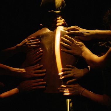 A person is seen from behind, waist up, illuminated by warm, subtle light sharply contrasted by shadow. A stripe of light runs along their spine, and multiple hands from outside the frame reach toward and touch them.