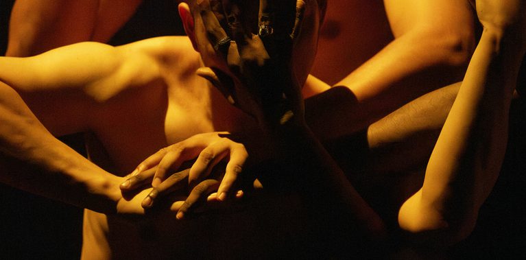 Two shirtless people are shown in a close-up from the waist up, illuminated by warm light sharply contrasted by dark shadow. The person behind covers their face with one hand and intertwines the other with the hand of the person in front. The front person’s arm is raised to chest level, elbow bent, and intertwined with the hand of another person to the left, who is outside the frame. Their face is also covered by the hand of someone unseen outside the frame.
