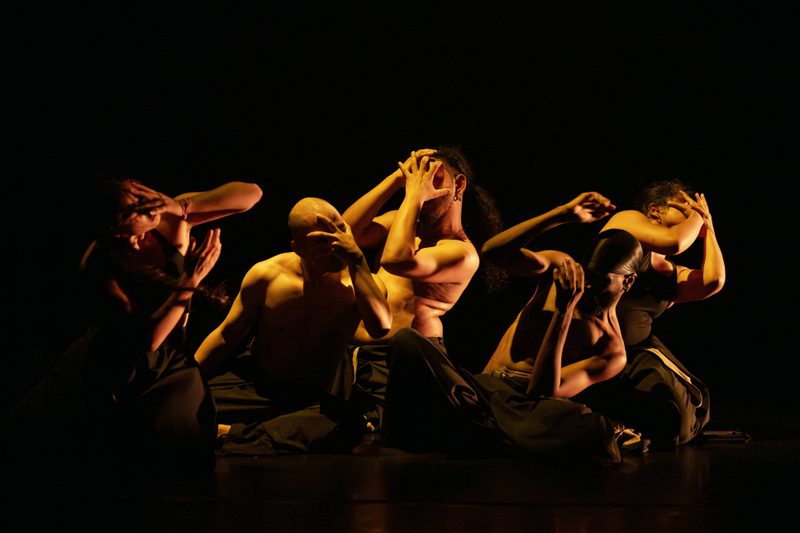 A group of people wearing black trousers—some with black tops, others shirtless—are kneeling and covering their faces with their hands, illuminated by warm light that contrasts with the surrounding darkness.