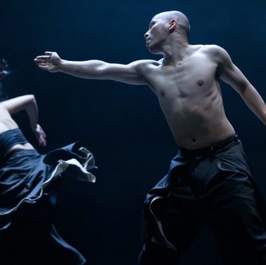 A person wearing black trousers, shirtless and bald, looks to the left and stretches their arm in that direction toward another person in a black top and skirt, caught mid-turn.