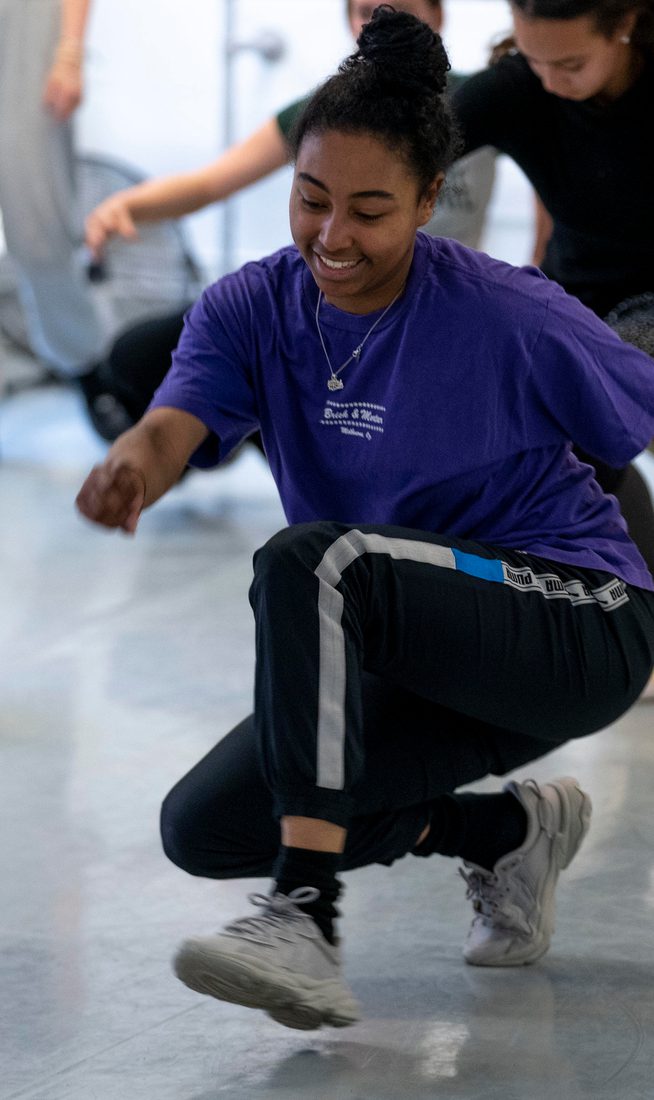 A teenager wearing a purple t-shirt and tracksuit bottoms in a dance studio crouching down and walking, there are several other teenagers in the studio behind them doing the same thing.