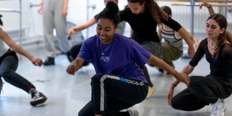 A teenager wearing a purple t-shirt and tracksuit bottoms in a dance studio crouching down and walking, there are several other teenagers in the studio behind them doing the same thing.