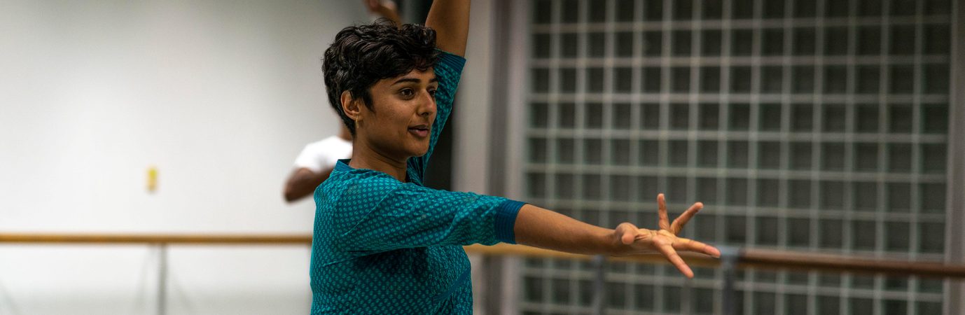 Bharatanatyam teacher, Shivaangee Agrawal, wears a turquoise top and has short black hair. Their right arm is completely outstretched in front of their body, palm facing up and fingers splayed. Their left are is above their head, two fingers and thumb pressed together in a classical mudra position.