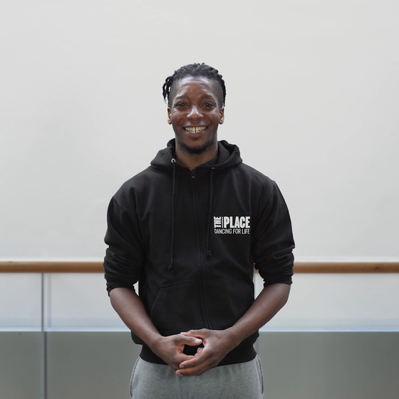 Person standing in a dance studio wearing a black hoody with The Place logo printed on it