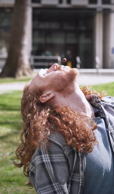 A person with long ginger hair sitting on some grass. They are leaning their head back and blowing a Malteaser in the air with their mouth.