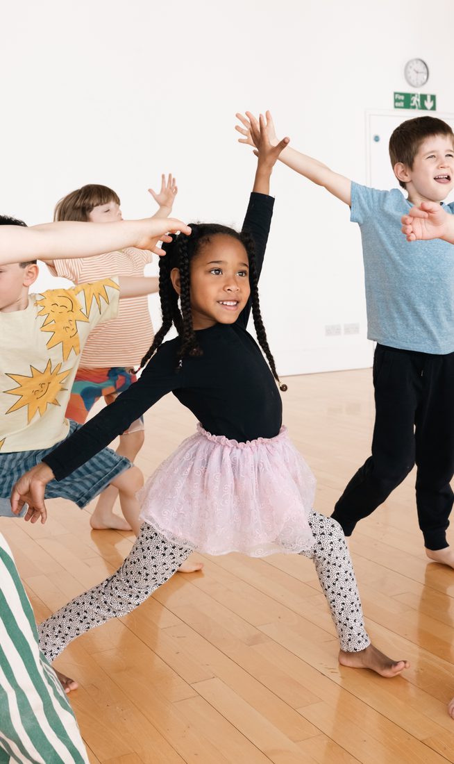 A girl around 5 years old dancing with her arms stretched out, wearing a black top and pink tutu.