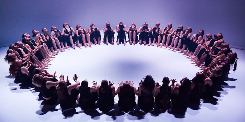 A large group of dancers, dressed in black sleeveless tops and trousers, are seated on a white floor in a perfect circle. They are all leaning back slightly with their hands raised in front of them, palms facing inward toward the center of the ring. The scene is captured from a high angle under cool, blue-toned lighting that casts soft shadows.