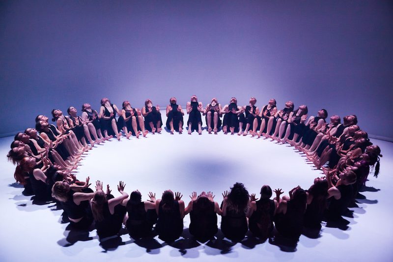 A large group of dancers, dressed in black sleeveless tops and trousers, are seated on a white floor in a perfect circle. They are all leaning back slightly with their hands raised in front of them, palms facing inward toward the center of the ring. The scene is captured from a high angle under cool, blue-toned lighting that casts soft shadows.