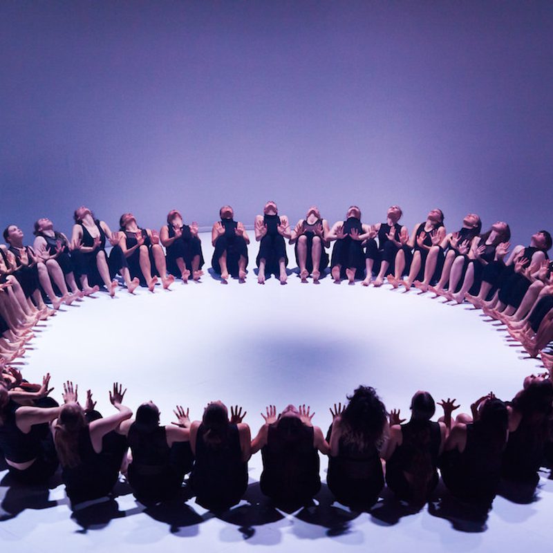 A large group of dancers, dressed in black sleeveless tops and trousers, are seated on a white floor in a perfect circle. They are all leaning back slightly with their hands raised in front of them, palms facing inward toward the center of the ring. The scene is captured from a high angle under cool, blue-toned lighting that casts soft shadows.