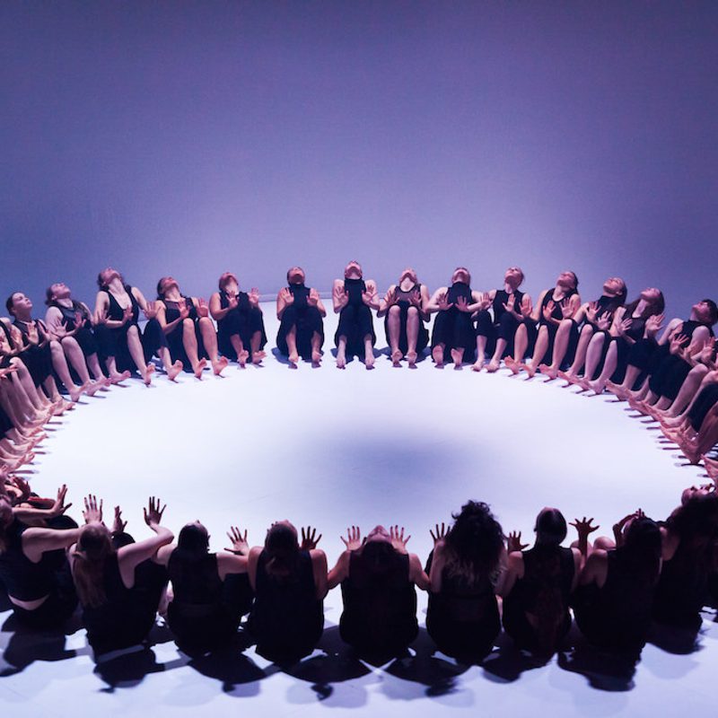 A large group of dancers, dressed in black sleeveless tops and trousers, are seated on a white floor in a perfect circle. They are all leaning back slightly with their hands raised in front of them, palms facing inward toward the center of the ring. The scene is captured from a high angle under cool, blue-toned lighting that casts soft shadows.