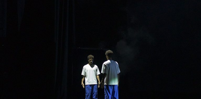 A lone performer stands on a dark, expansive stage in front of a tall mirror, facing their own reflection. They are wearing a white t-shirt and bright blue trousers. A single, sharp spotlight cuts through the darkness from above, illuminating the figure and creating a long, clear reflection on the polished black floor. Faint wisps of stage smoke are visible in the beam of light.