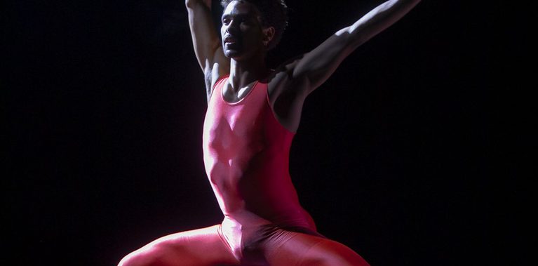 A solo dancer in a bright, neon-red unitard performs on a dark stage. He is captured in a wide, low second-position lunge with his arms extended dynamically – one reaching straight upward and the other out to the side. A cool-toned spotlight hits his body from the side, creating sharp highlights against the black background and reflecting off the polished stage floor.