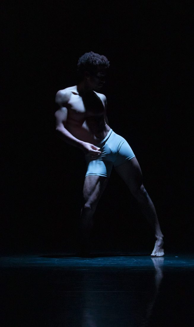 A dancer wearing light-colored briefs performs a solo on a pitch-black stage. He is captured in a dynamic, leaning pose with one leg extended to the side and his torso twisted slightly. A cool, blue-toned spotlight illuminates his physique from the side, casting deep shadows and highlighting his muscle definition, while his reflection is faintly visible on the stage floor.
