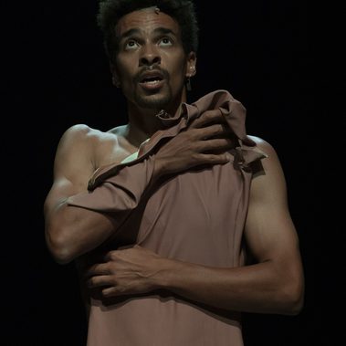 A close-up of a performer looking upward with an open-mouthed, emotive expression. He is shirtless on one side, clutching a draped pink fabric across his chest with both hands against a dark, minimalist background.