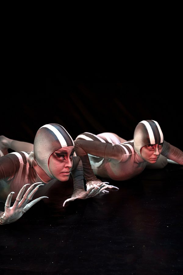 Two people caught in a prowling motion in a dark theatre setting, wearing skintight grey-and-white costumes with matching caps, makeup, and gloves tipped with long, spiky fingers, evoking lizard-like creatures.