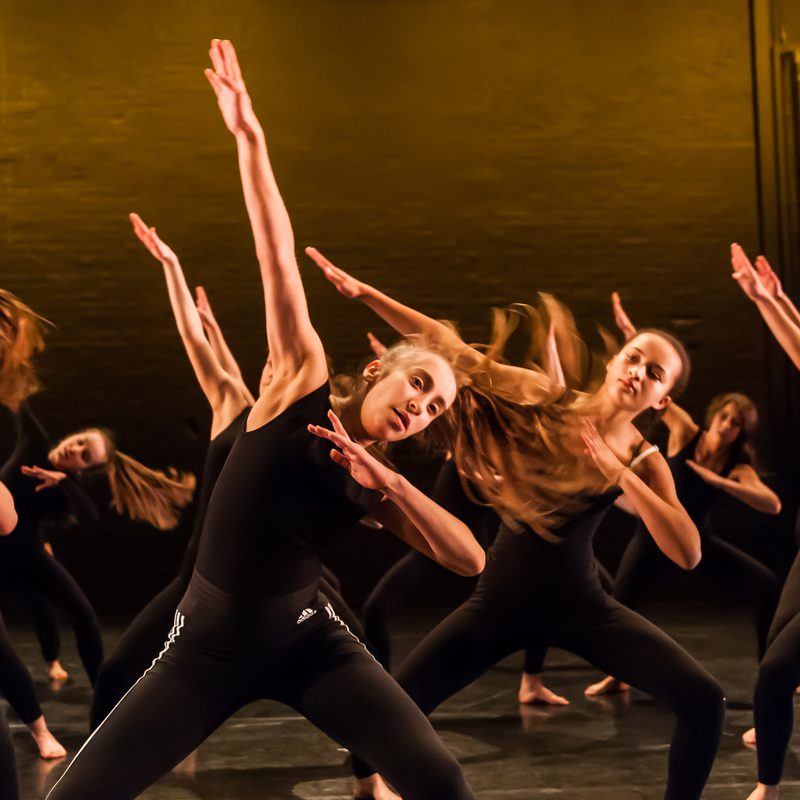 A group of secondary school students on The Place stage wearing black tops and trousers dancing