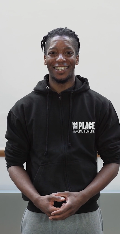 A person standing in a dance studio with a ballet barre behind them. They are wearing a black hoody with The Place logo on in the top right corner