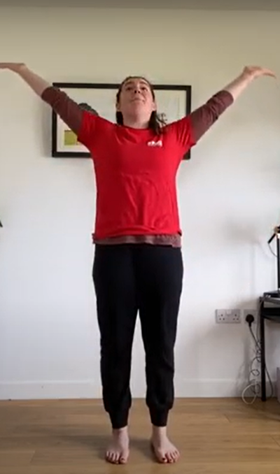 Dance teacher in their lounge wearing a red t-shirt and black trousers stretching their arms upwards towards the ceiling.