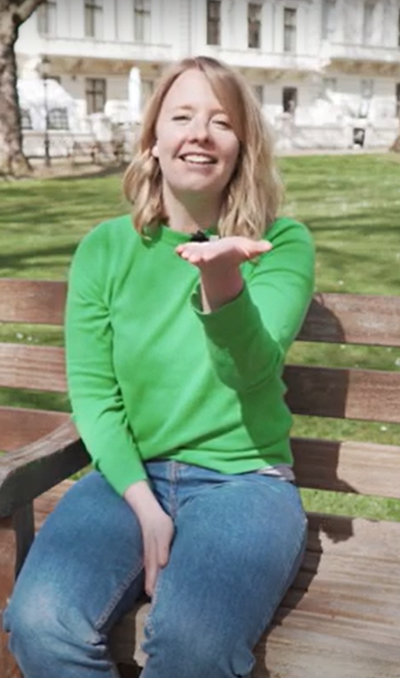 Person sitting on a bench outside wearing a green jumper and jeans. They are holding out their hand palm faced up and smiling.