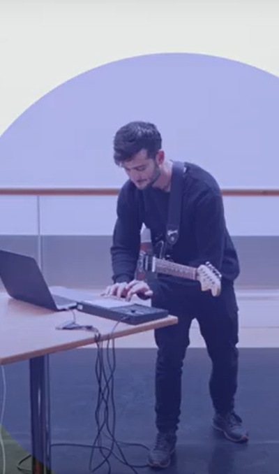 Person with a guitar in a dance studio taps into a keyboard. There is a blue opaque circle round them.
