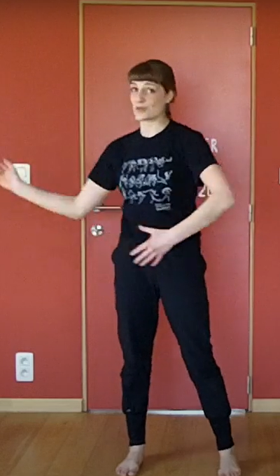 Person in a room with a red wall and wooden floor. They are wearing a black t-shirt and black trousers and are moving their arms in front of them.