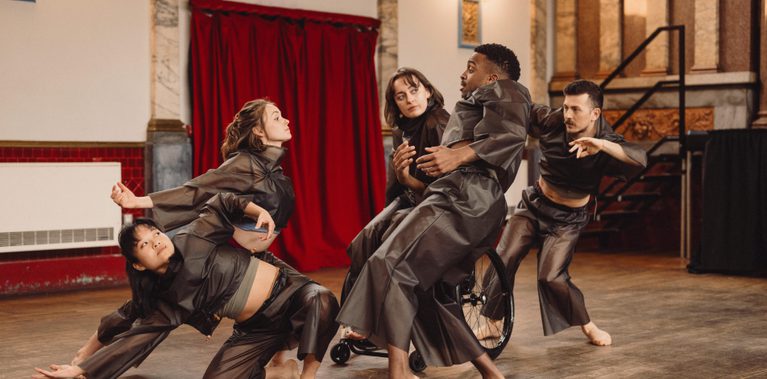 Five dancers are positioned in an ornate hall with marble columns and a red velvet backdrop. They wear dark, translucent oversized tracksuits. In the center, a dancer in a wheelchair is tilted backward while being supported by a standing performer. The other three dancers are captured in mid-movement, crouching and lunging at different levels across the wooden floor.