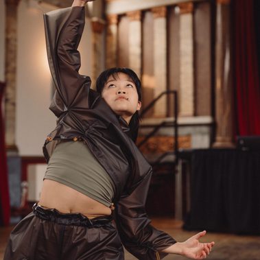 A close-up of a dancer in a translucent, dark oversized tracksuit over a green crop top. They are captured mid-movement, reaching one arm high with a clenched fist while looking upward. The background features marble columns and architectural details of the hall.
