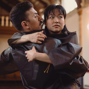 A close-up shot of two performers in a space with a high, vaulted ceiling. They are wearing dark, oversized matte tracksuits. One performer has their back to the camera and is being held by the second performer, who looks off into the distance with a serious, contemplative expression. The lighting is warm and natural, emphasizing the texture of their clothing and the emotion of the moment.