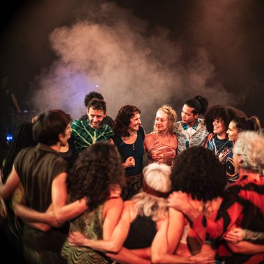 A diverse group people standing in a circle, smiling, about to go on stage. They are wearing colourful costumes that resemble vegetation. Behind them is smoke and the colours of stage lighting.