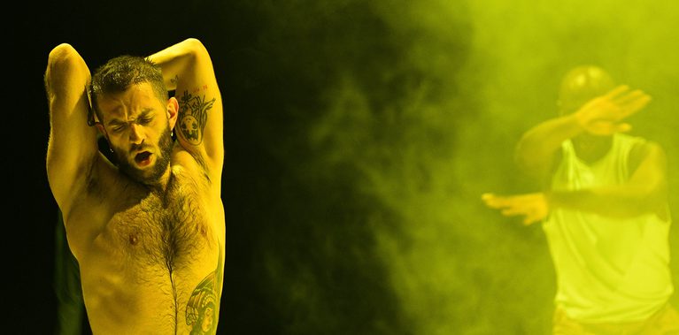 A performer stretches with their arms behind their head under yellow stage lighting, while a second performer moves in the background through haze.