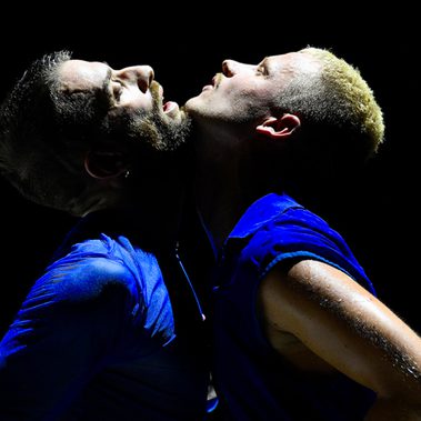 Two performers in blue costumes stand back-to-back under a spotlight on a dark stage.