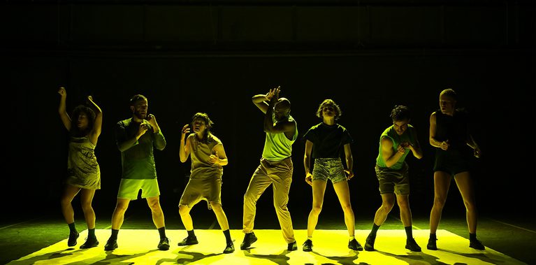 Seven performers stand in a line on a bright yellow-lit stage, each holding a different pose with bent arms, raised hands or angled torsos against a dark backdrop.