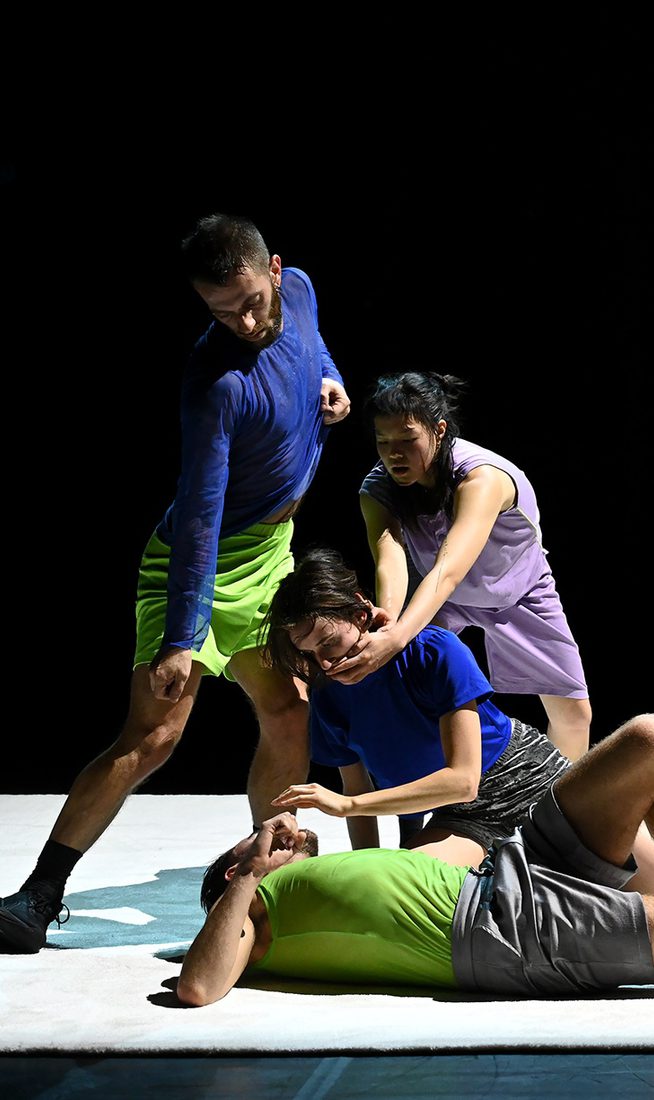 Seven dancers perform together on a white stage against a black background. Their bodies form a clustered composition of standing, crouching, kneeling, and reclining positions, with several performers making physical contact. Bright blue, green, and purple costumes stand out under stage lighting.