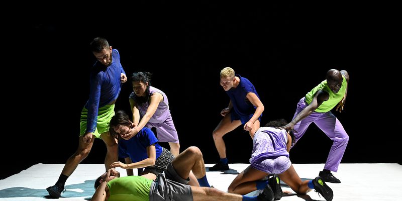 Seven dancers perform together on a white stage against a black background. Their bodies form a clustered composition of standing, crouching, kneeling, and reclining positions, with several performers making physical contact. Bright blue, green, and purple costumes stand out under stage lighting.