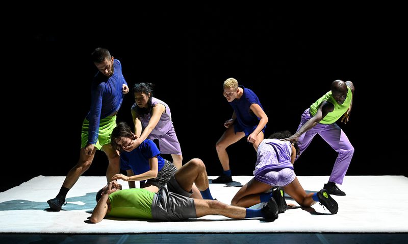 Seven dancers perform together on a white stage against a black background. Their bodies form a clustered composition of standing, crouching, kneeling, and reclining positions, with several performers making physical contact. Bright blue, green, and purple costumes stand out under stage lighting.