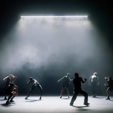 Six performers are dispersed across a dimly lit stage, crouching, leaning, and gesturing beneath a bright overhead light that illuminates the haze above them.
