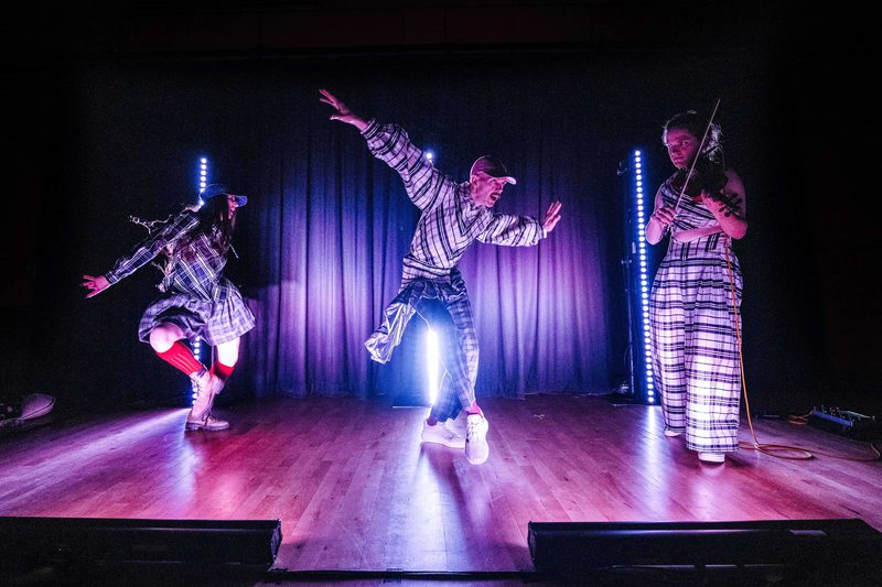 Three figures dressed in tartan on a pink and purple lit stage