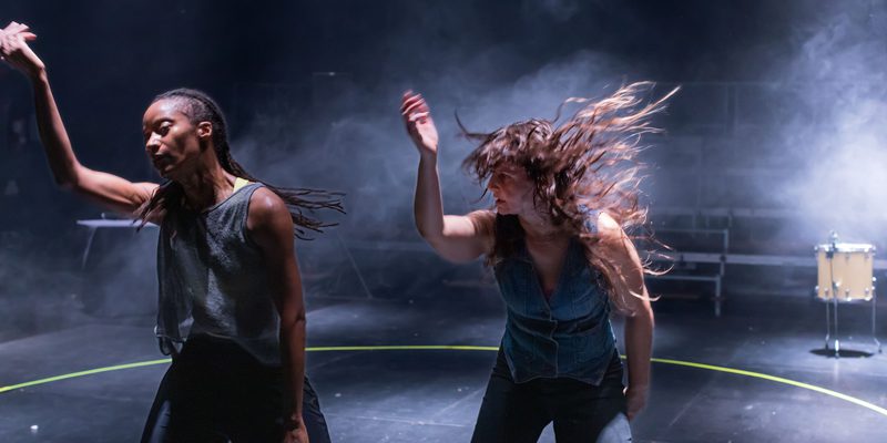 Two dancers perform inside a circular performance area on a dimly lit stage. Both raise one arm in mid-motion, while one dancer's hair is caught in motion. Haze and stage lighting fill the background, and a drum is visible at the edge of the space.