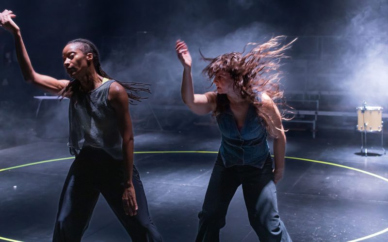 Two dancers perform inside a circular performance area on a dimly lit stage. Both raise one arm in mid-motion, while one dancer's hair is caught in motion. Haze and stage lighting fill the background, and a drum is visible at the edge of the space.
