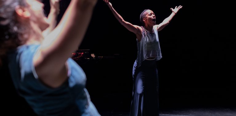 Two dancers perform on a dark stage under spotlights. In the foreground, a blurred figure raises their arms overhead, while a second dancer in the background stands with both arms outstretched and their mouth open. A curved line marking the performance area is visible on the floor.