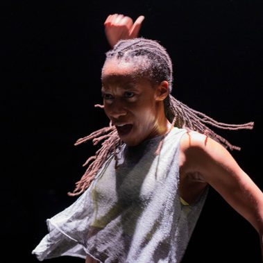 Close-up of a dancer in motion under stage lighting. Their braided hair swings outward as one arm lifts above their head and the other extends to the side. The dancer's face is animated, and the dark background isolates the movement.