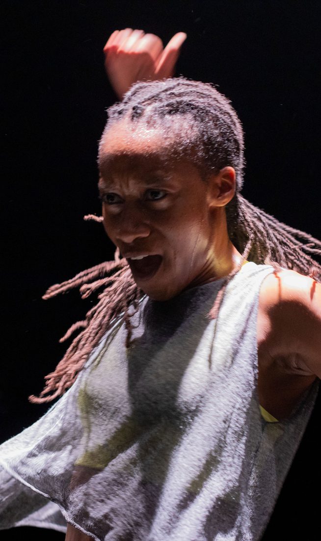 Close-up of a dancer in motion under stage lighting. Their braided hair swings outward as one arm lifts above their head and the other extends to the side. The dancer's face is animated, and the dark background isolates the movement.