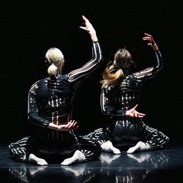 Two figures wearing striped body suits, face away from the camera, with one arm raised, the other behind their backs
