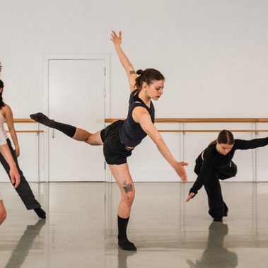 A group of students are warming up in a studio, the student in the centre raises one leg high behind them with their arms parallel