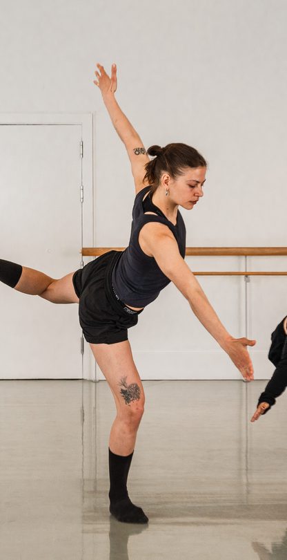 A group of students are warming up in a studio, the student in the centre raises one leg high behind them with their arms parallel